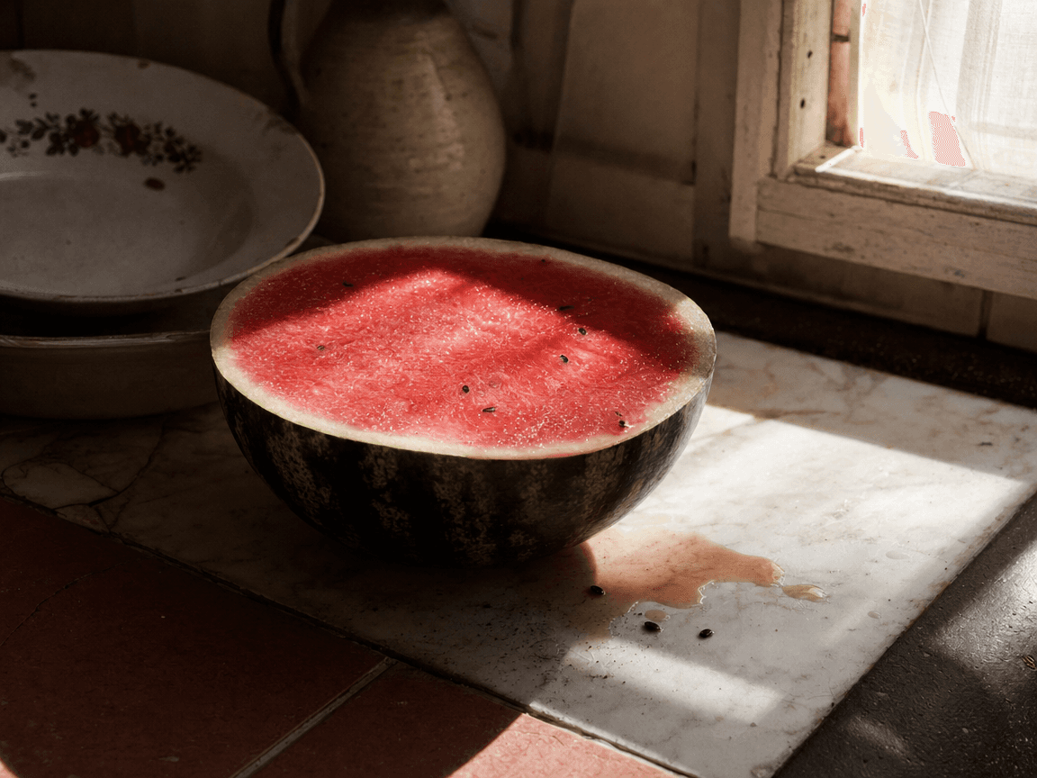 Une pastèque coupée en deux posée sur une table de cuisine en carrelage ancien, la chair rouge vif luisant sous la lumière rasante d'une fenêtre d'été, quelques graines noires et une flaque de jus sur le marbre.