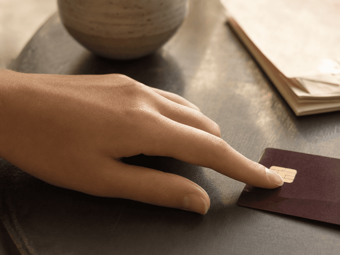 Une main aux doigts légèrement flous posée à plat sur une table en bois sombre, l'index effleurant le capteur doré d'une carte bancaire, dans une lumière de fin d'après-midi qui rasante fait apparaître les crêtes et les sillons absents de la peau.