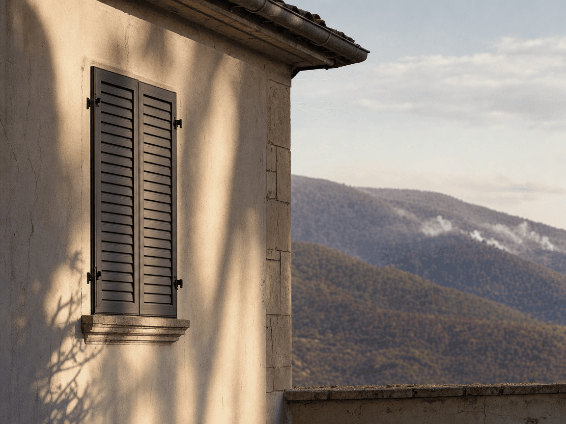 Une façade de maison en enduit minéral clair, léchée par une lumière rasante de fin d'après-midi, ses volets métalliques fermés se détachant sur un fond de collines roussies et encore fumantes.