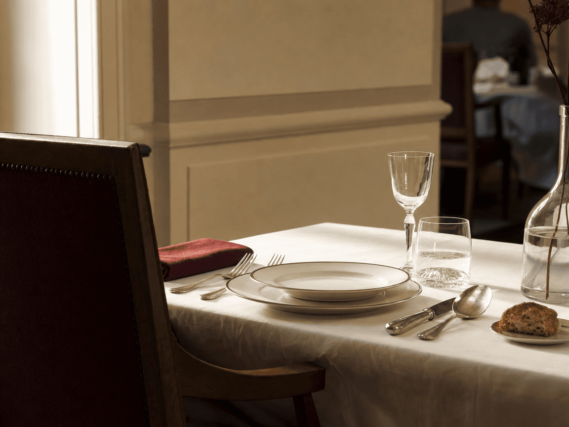 Une table de salle à manger commune dressée pour personne, couverts alignés avec soin sur nappe blanche légèrement froissée, lumière de fin de matinée rasant les verres vides.