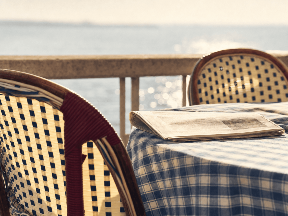 Une terrasse de café en bord de mer, tables encore vides et chaises en osier tournées vers la lumière rasante d'un matin de septembre, un journal plié posé sur la nappe à carreaux bleus.